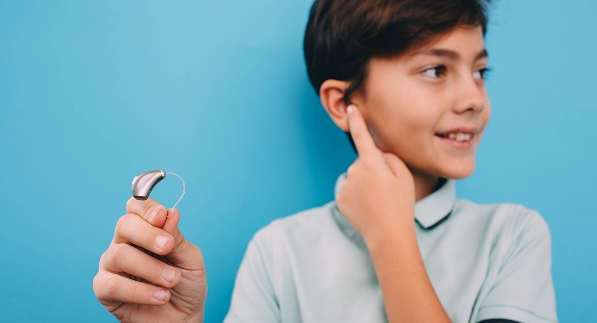 Kind hält Hörgerät in der Hand und zeigt auf sein Ohr – moderne Kinderhörgeräte für den Schulalltag