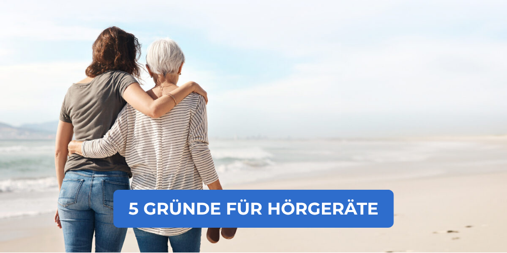 Erwachsene Tochter und ältere Mutter gehen Arm in Arm am Strand entlang und blicken aufs Meer – 5 Gründe, warum sich Hörgeräte lohnen