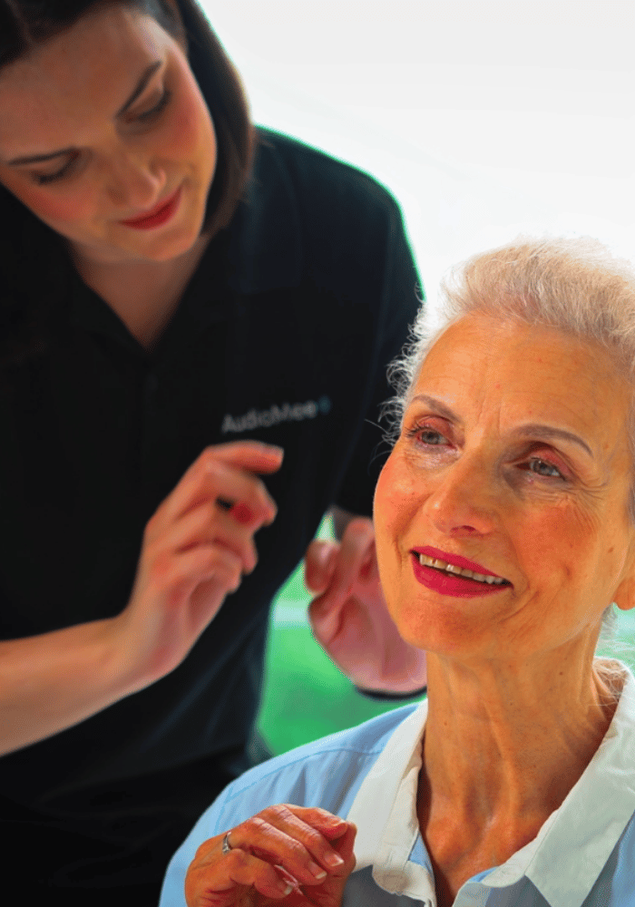 AudioMee Hörakustikerin passt einer lächelnden Kundin ein Hörgerät an – Probetragen und Auswahl bei der Erstversorgung zu Hause